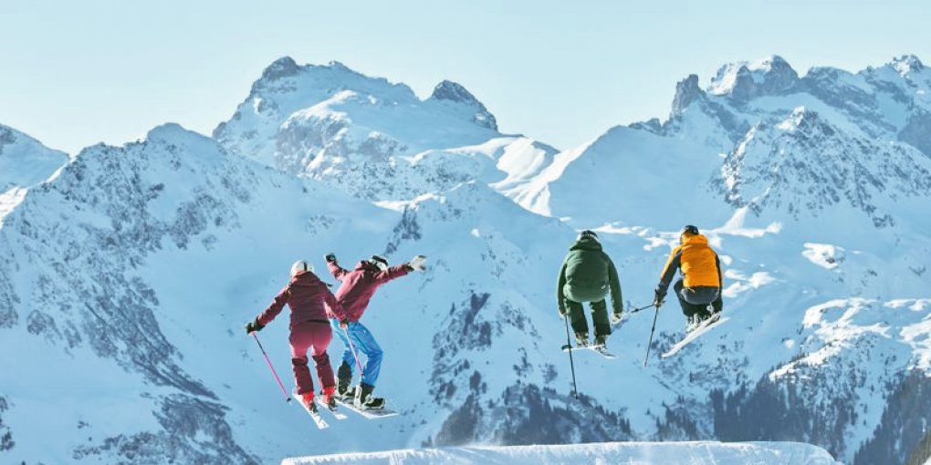 Vier Jugendliche schanzen mit Skiern und Snowboard über Schanze vor einer schneebedeckten Bergkulisse. © Silvretta-Montafon - Stefan Kothner