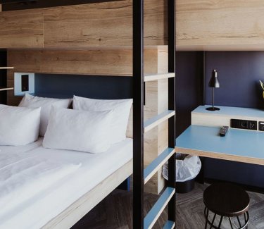 Doppelbett mit weißem Bezug, darüber Stockbett mit Leiter, daneben kleiner Schreibtisch mit Lampe und Hocker. © schulz hotels