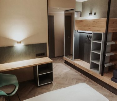 In Brauntönen gehaltene Möbel: links Schreibtischmit Stuhl, daneben Stockbett mit offenem Schrank darunter und Spiegel, rechts Sofa. © Schulz Hotels