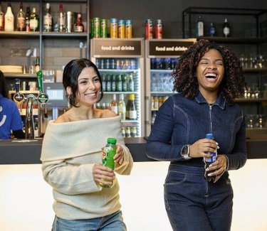 Zwei junge Frauen stehen vor einer Bartheke mit einer Flasche in der Hand und lachen herzhaft. © a&o hostels
