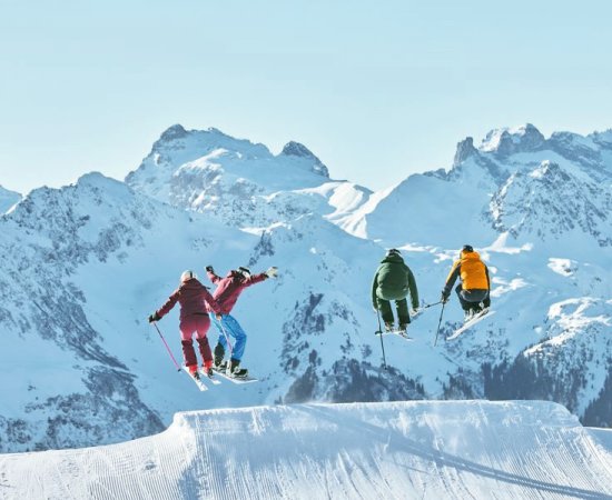Vier Jugendliche schanzen mit Skiern und Snowboard über Schanze vor einer schneebedeckten Bergkulisse. © Silvretta-Montafon - Stefan Kothner